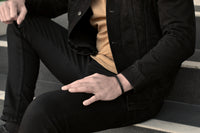 Sitting man in casual black jeans outfit wears a sleek black leather bracelet from Rianrix Accessories Parker - Single-layer leather bracelet with fishbone pattern, fashion picture close up