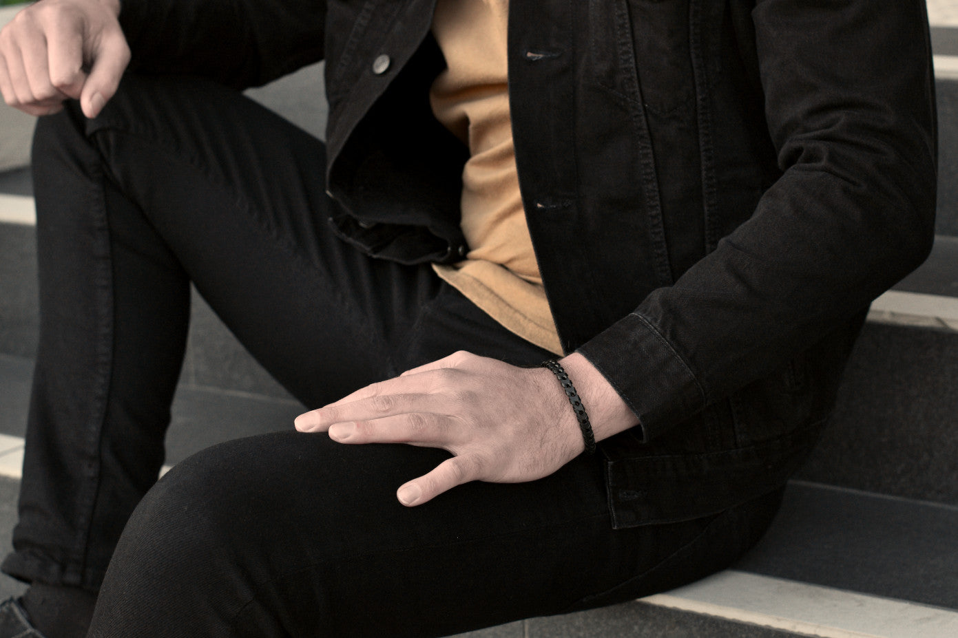 Sitting man in casual black jeans outfit wears a sleek black leather bracelet from Rianrix Accessories Parker - Single-layer leather bracelet with fishbone pattern, fashion picture close up