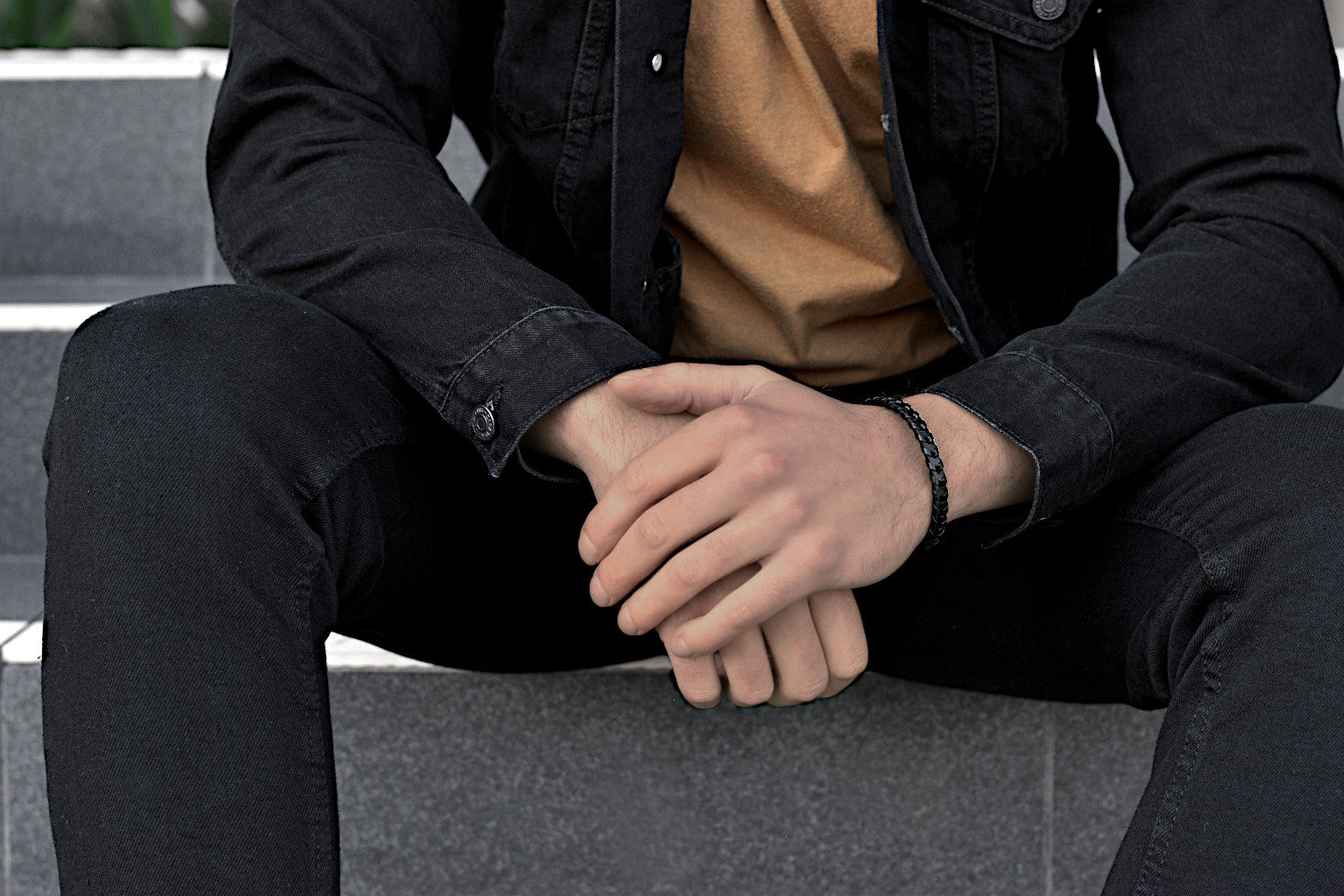 Sitting man in casual black jeans outfit wears a sleek black leather bracelet from Rianrix Accessories 